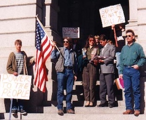 A group of around eight people stand on the steps of the Colorado capitol holding signs and a flag.