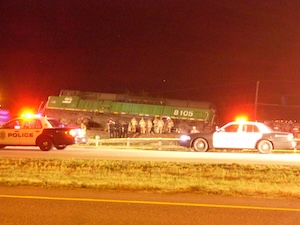 Derailed train engine with emergency workers and flashing patrol cars in front.