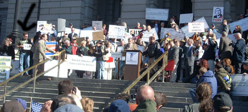 A crowd gathers atop the front steps of the Colorado Capitol. An arrow points to a man holding a sign on the left edge of the crowd.