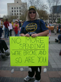 A woman holds a sign reading No More Spending We're Broke and So Are You.