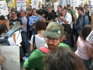 A group of people and journalists gather, one person holds a sign reading Pro Muslim Anti Racist socialistworker dot org.