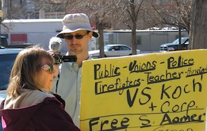 Ari Armstrong holding a video camera, while a woman in front holds a sign reading public unions police firefighters teacher principles vs Koch and corp.