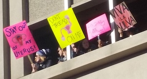 Several people peer down from a window, including a woman holding a sign reading stay out of my uterus.