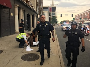Two Denver police officers approach Susan Greene while other officers converge around a man handcuffed on the ground.