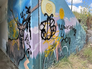 A once-beautiful mural on the side of a road underpass is vandalized with ugly spray paint.
