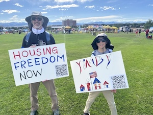 At the July 4, 2025, celebration in Westminster, Ari Armstrong holds a sign saying 'Housing Freedom Now,' while his son holds a YIMBY sign. A scan code links to a sign-up form for YIMBY North Metro.